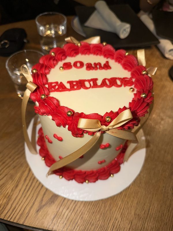 A white cake with red piped borders, gold bows and a 50 and Fabulous topper