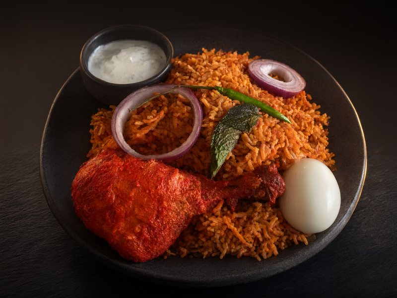 Chilli chicken biryani on a dark plate with a red tandoori chicken leg, a boiled egg, red onion rings, a green chilli, mint and a bowl of raita